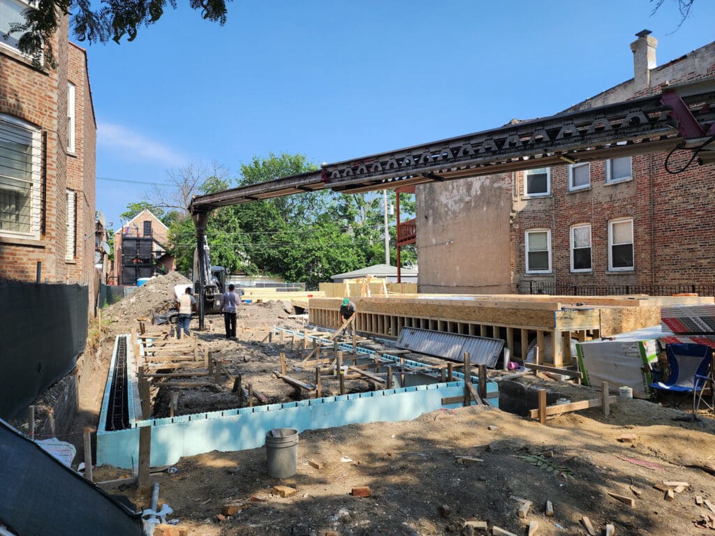 Construction site with workers operating machinery and laying foundation surrounded by brick buildings and trees under clear blue sky.